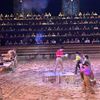 Performers are engaged in an action-packed scene in a sandy arena, while a captivated audience watches from tiered seating in the background.