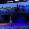 The image depicts a dimly-lit theatrical scene with performers dressed in Native American-inspired costumes on a stage that resembles a rugged landscape, with teepees projected in the background.