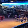 The image shows a theatrical performance with performers in colorful medieval-style costumes and a horse-drawn carriage inside an arena with a backdrop that resembles a rocky landscape.