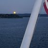 A full moon rises over a tranquil sea, viewed from the deck of a boat with a glimpse of its flagpole in the foreground.