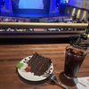 A slice of chocolate cake and a glass of a dark drink, possibly soda, are on a table with a view of a theater stage in the background.