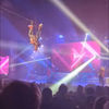 A performer is suspended mid-air during an aerial acrobatic act at a live event with colorful stage lighting and an audience in the foreground.