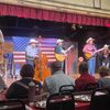 A band of musicians wearing cowboy hats is performing on stage in front of an audience with an American flag as the backdrop.