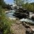 The image depicts a serene artificial river with cascading waters flanked by rocky banks, lush greenery, and rustic structures, possibly a part of an amusement park or themed attraction.