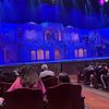 The image depicts a theater auditorium with an ornately decorated stage set for a production, with audience members seated and waiting for the performance to begin under a lighting setup that casts a blue hue over the scene.