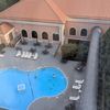 The image shows a bird's-eye view of a kidney-shaped swimming pool with a few people in it, surrounded by lounge chairs and a hotel-like building with an open patio area, taken from behind a window or barrier.