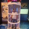 A person is standing inside a transparent hurricane simulator machine with a display showing a wind speed of 78.9 MPH above them.