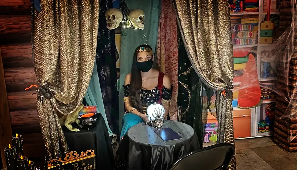 A person dressed in a fortune teller or mystical themed costume sits behind a table with a crystal ball in a tent-like setup with skull decorations and dim lighting