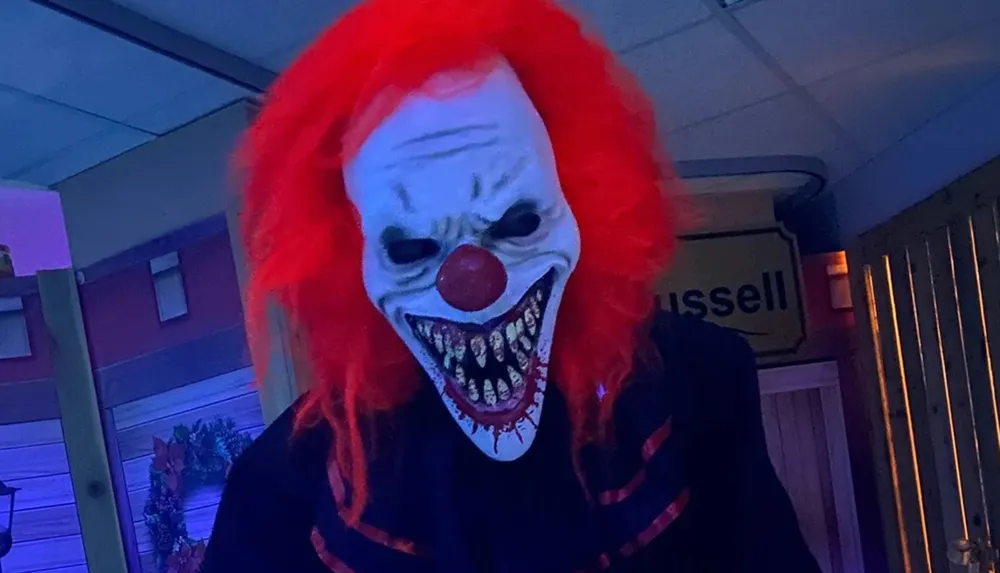 A person is wearing a scary clown mask with bright red hair and a wide menacing grin standing indoors under a blue light