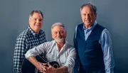 Three smiling men, one holding a guitar, pose together for a friendly portrait against a gray background.