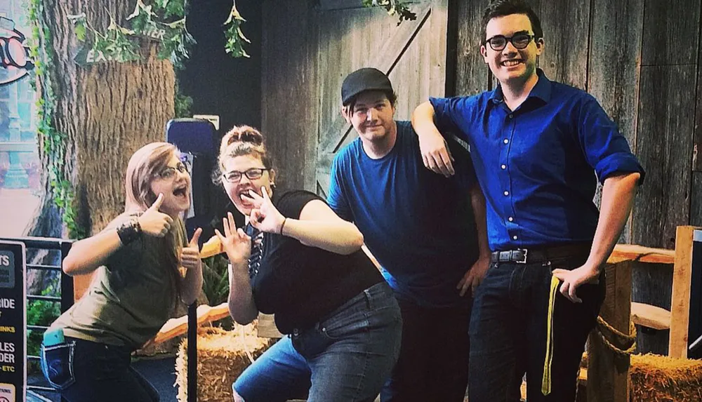 Four individuals are posing playfully for a photo in front of a rustic wooded backdrop with two of them making hand gestures that suggest a rock and roll or playful vibe