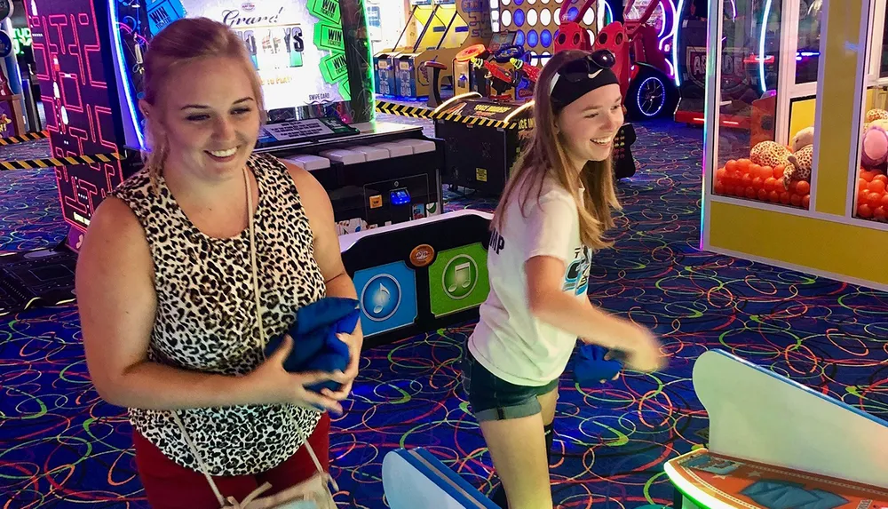 Two people are enjoying themselves in a vibrant arcade with one person playing a game and the other watching and smiling