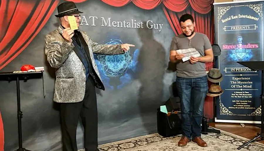 A person in a shiny silver jacket and hat is performing on stage, pointing at a smiling audience member who is reading something, both surrounded by mentalism-themed decor.