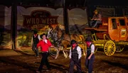 Performers in Wild West attire are present in front of a stagecoach, with one man smiling and twirling a lasso, evoking the atmosphere of a classic western show.