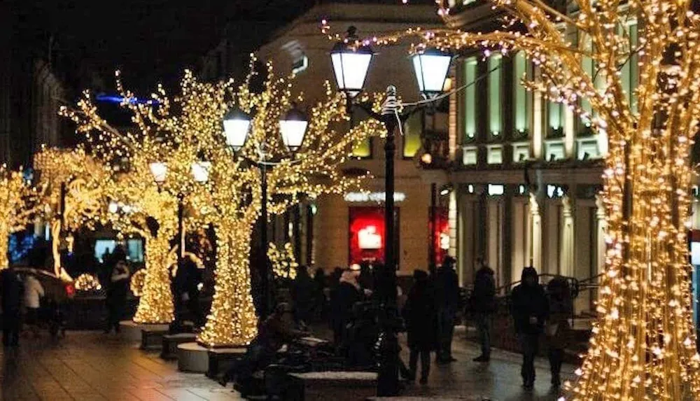 The image shows a festive evening scene on a street lined with trees illuminated with golden fairy lights as people walk by and enjoy the ambiance