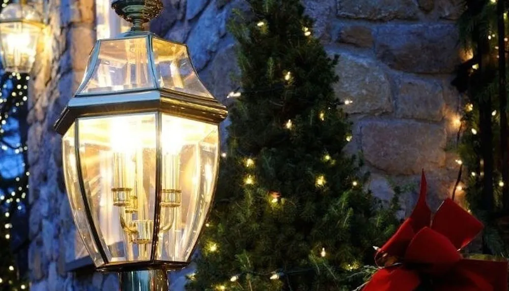 A classic street lamp is illuminated against a backdrop of a stone wall and tree decorated with twinkle lights and a red bow evoking a cozy holiday ambiance