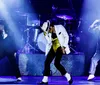 Three performers onstage exhibit a dynamic dance pose reminiscent of Michael Jacksons iconic style with the central figure donning a white jacket and gold belt