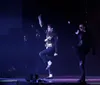 Three performers onstage exhibit a dynamic dance pose reminiscent of Michael Jacksons iconic style with the central figure donning a white jacket and gold belt