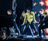 Three performers onstage exhibit a dynamic dance pose reminiscent of Michael Jacksons iconic style with the central figure donning a white jacket and gold belt