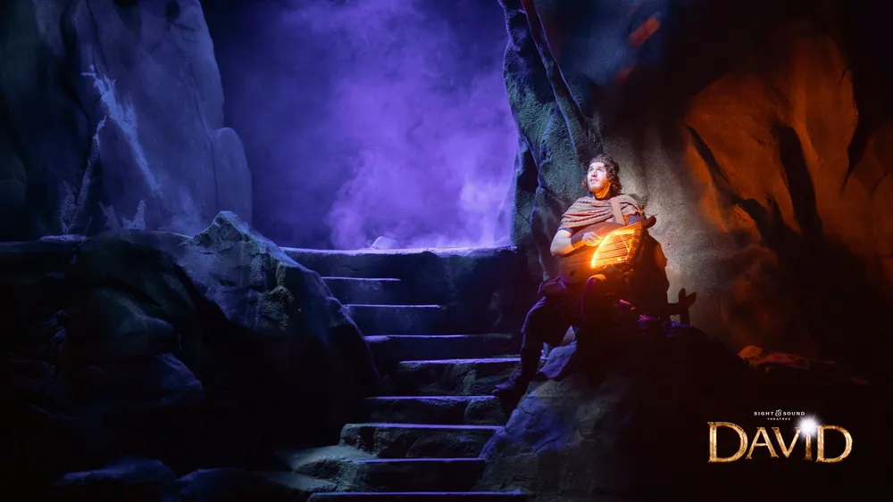 A person in historical-looking attire sits on stone steps in a cavernous environment illuminated by the warm glow of a lantern with purple-hued fog in the background and the word DAVID displayed prominently at the bottom