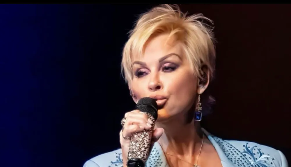 A woman with a pixie haircut is singing into a bejeweled microphone wearing a blue outfit with decorative details