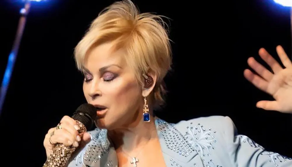 A performer with short blonde hair is singing into a microphone wearing a blue embellished outfit and expressive makeup with their hand gesturing mid-performance