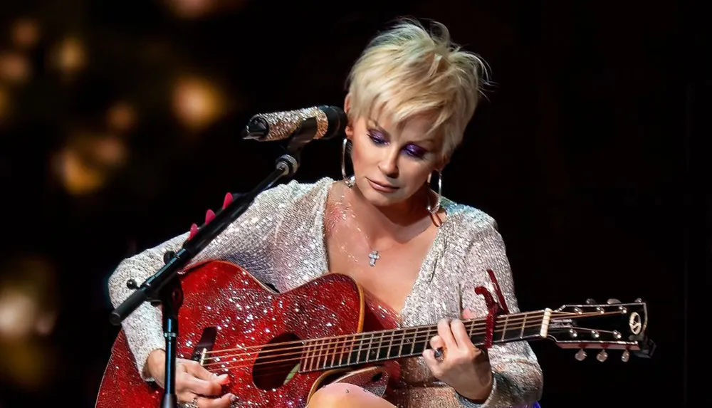 A person with a short blonde haircut is performing on stage playing a red glittery guitar and wearing a sparkly outfit