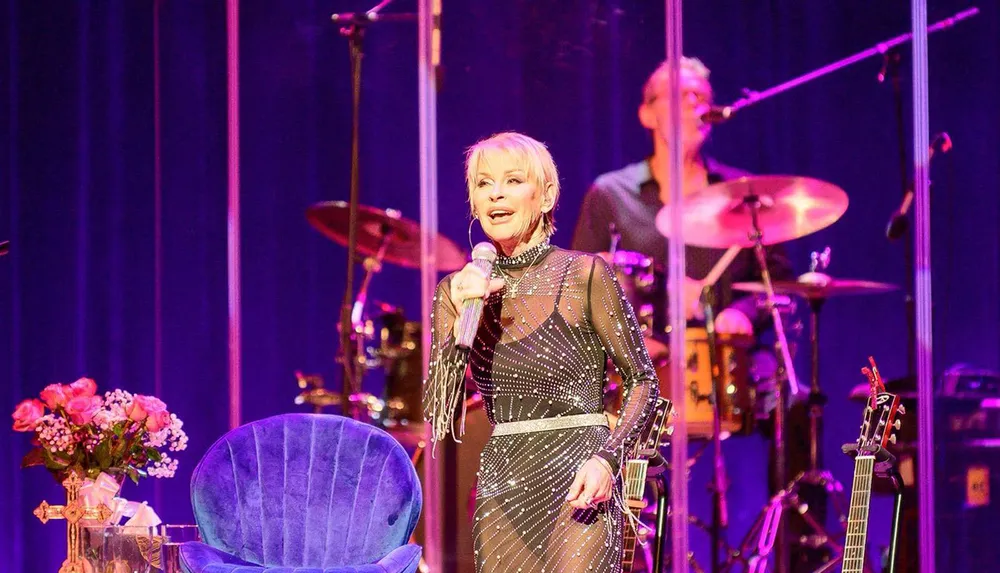 A performer is singing on stage with a band in the background amidst musical equipment and a bouquet of flowers on a stool