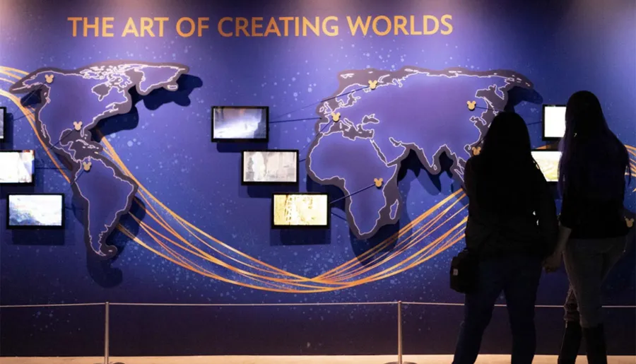 Two people are observing an exhibit titled THE ART OF CREATING WORLDS with world map graphics and multiple screens displaying content on a gallery wall.