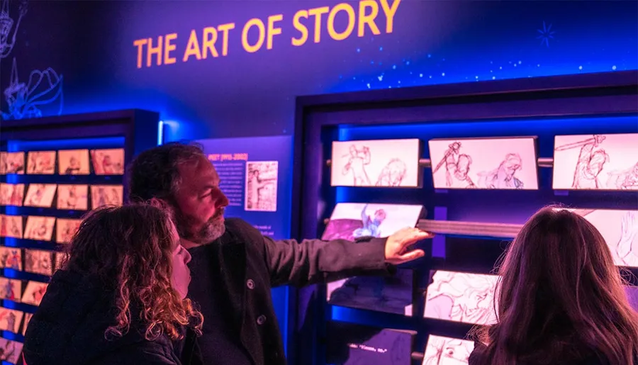 A person is showing something on a screen to two others in an exhibition space dedicated to THE ART OF STORY.