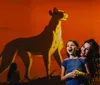 A woman and a young girl pose with bright smiles in front of a large projected animation of a cheetah against an orange backdrop