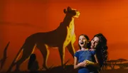 A woman and a young girl pose with bright smiles in front of a large, projected animation of a cheetah against an orange backdrop.