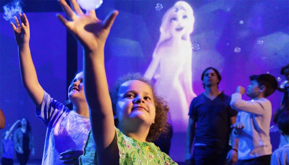 Children reach towards bubbles with joy in a room illuminated by a projection of an animated character