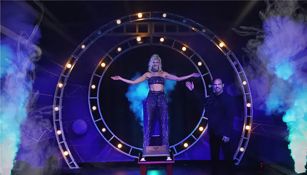 A magician poses next to an assistant standing inside a lit circular portal surrounded by theatrical smoke on stage