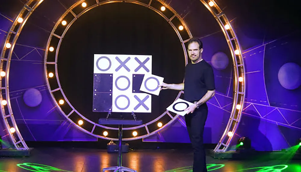 A person is standing on stage holding a O game piece in front of a large tic-tac-toe board with a circular light structure in the background