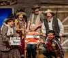 The image depicts a lively scene from a theatrical performance called Whodunnit Hoedown as a part of Branson Dinner Shows with actors dressed in western-style costumes expressing a range of exaggerated emotions