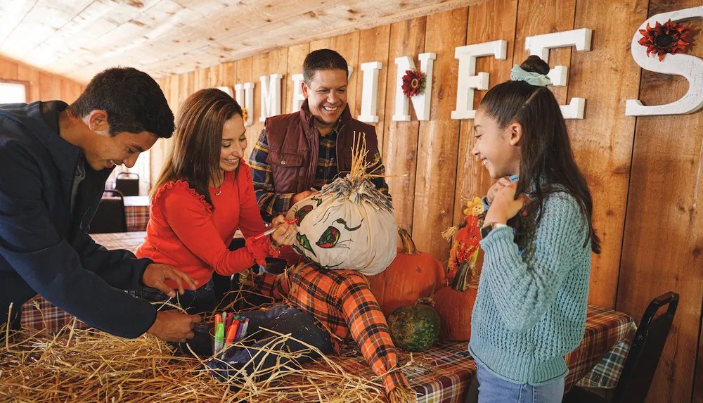 A family is joyfully decorating a scarecrow with colorful markers in a festive autumn-themed setting