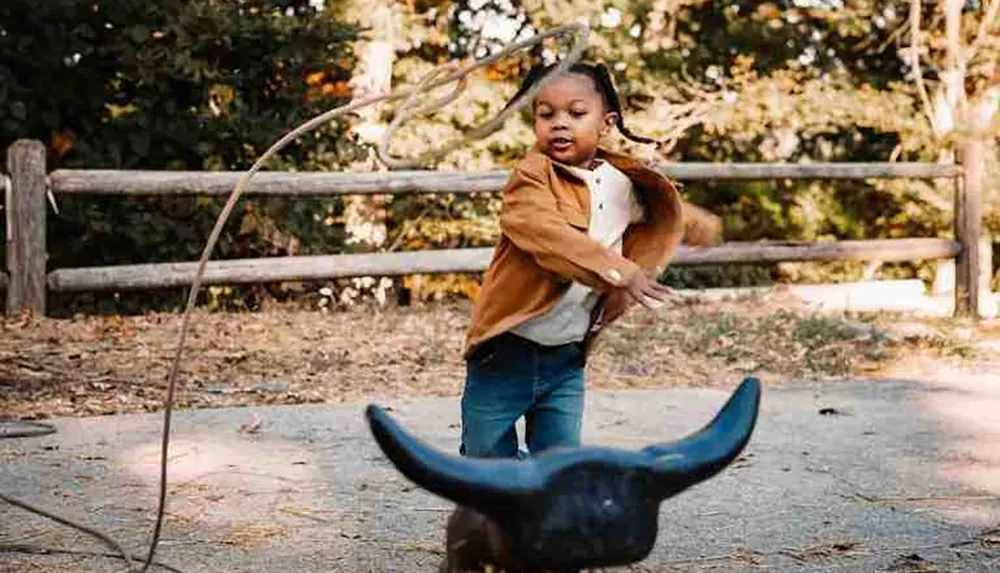 A child is playfully swinging at a plastic bull with a stick in an outdoor setting reminiscent of a mock bullfight