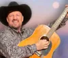 A smiling person in a cowboy hat holds a guitar up in the air against a soft-focus background