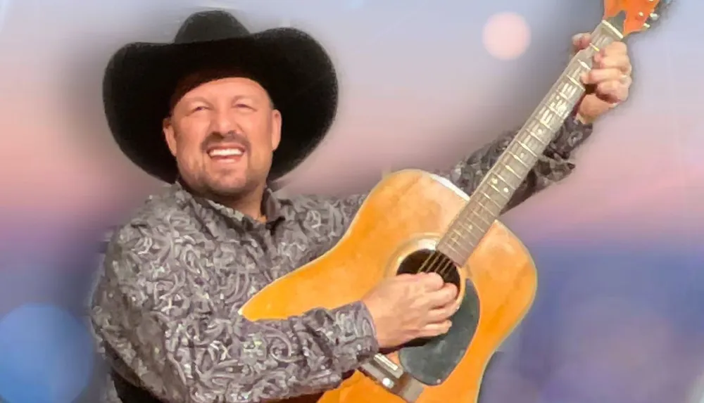 A smiling person in a cowboy hat holds a guitar up in the air against a soft-focus background