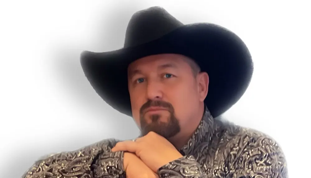 A man with a beard is wearing a black cowboy hat and a patterned shirt posing with crossed hands in front of a white background