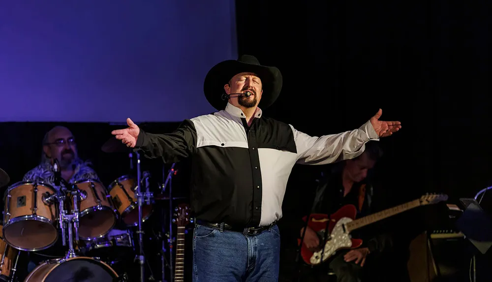 A man in a cowboy hat and a two-tone shirt passionately sings with a microphone headset on stage gesturing with his arms wide open as band members play in the background