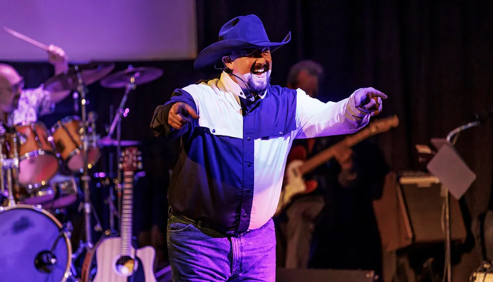 A man in a cowboy hat and a two-toned shirt is exuberantly performing on stage with a microphone in hand while a band plays instruments in the background