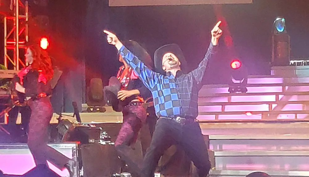 A person in a cowboy hat joyfully raises their arms while performing on a stage with bright lights and dancers in the background