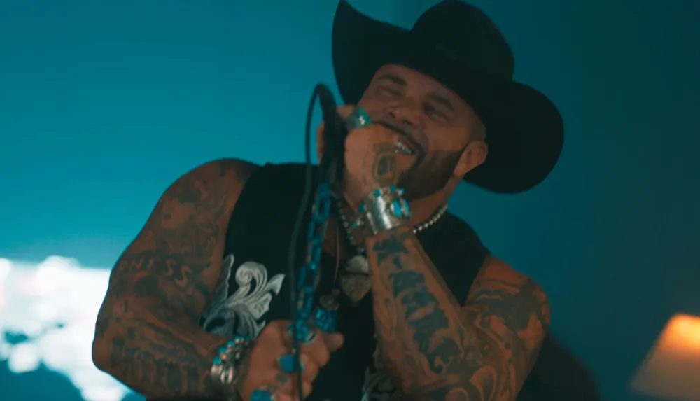 A tattooed man in a cowboy hat is singing passionately into a microphone with a joyful expression on his face