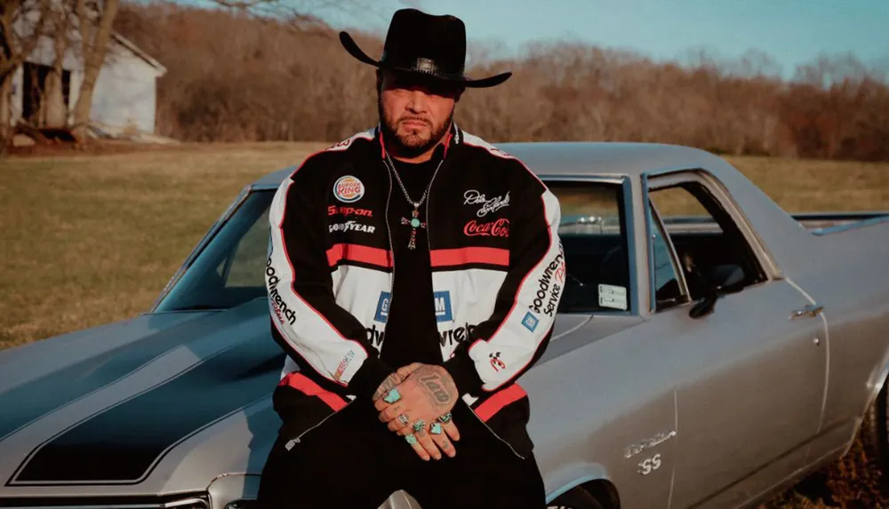 A man wearing a black hat and a racing jacket leans on a vintage muscle car in a rural setting