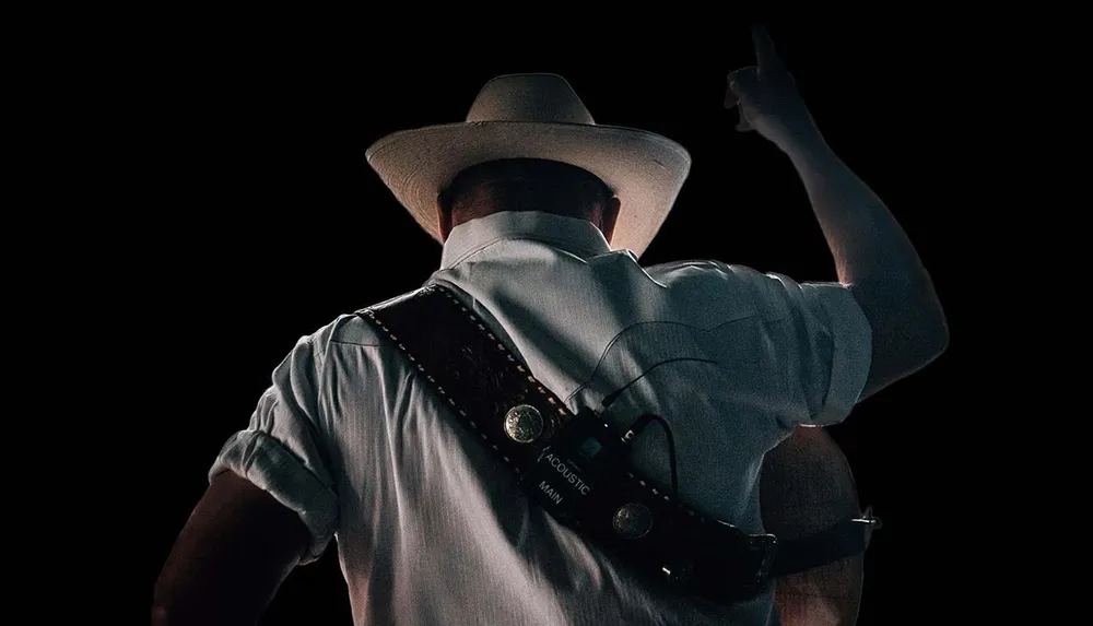 A person wearing a cowboy hat and a guitar strap is gesturing upwards with their finger against a dark background