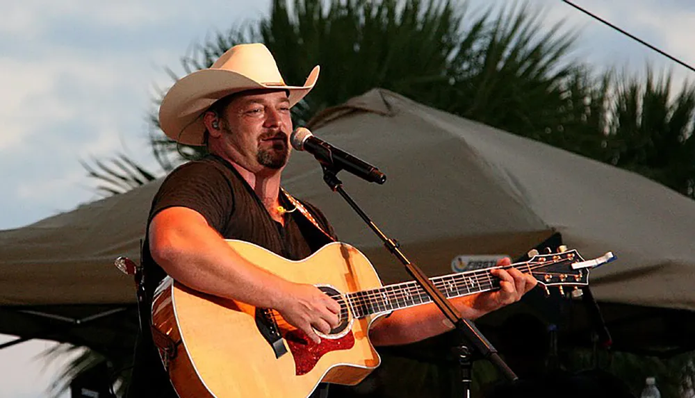 A man wearing a cowboy hat is playing an acoustic guitar and singing into a microphone on a stage with palm trees in the background