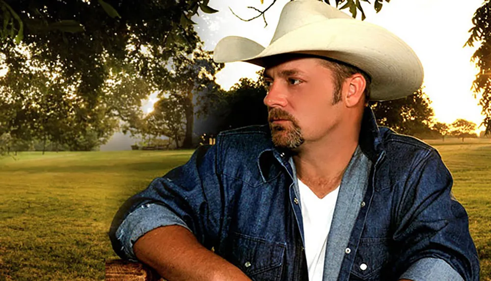 A man wearing a cowboy hat and a denim jacket is gazing into the distance in a pastoral scene with trees and a sunset