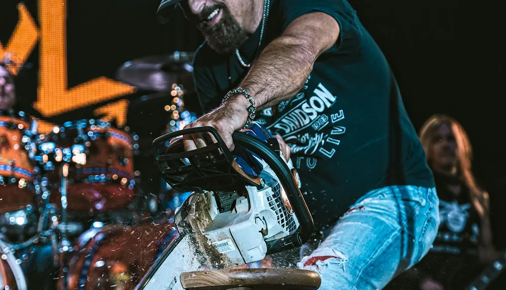 A person wearing jeans and a Harley-Davidson t-shirt is using a chainsaw on a wooden object with wood chips flying and a blurred drummer is visible in the background on stage